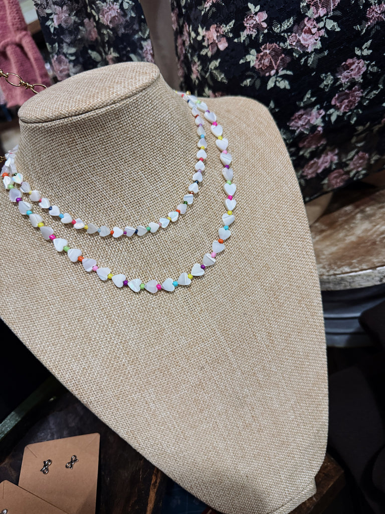 Two necklaces (one larger and one smaller) with heart-shaped beads and multicolor spacer beads on a mannequin bust with a floral background