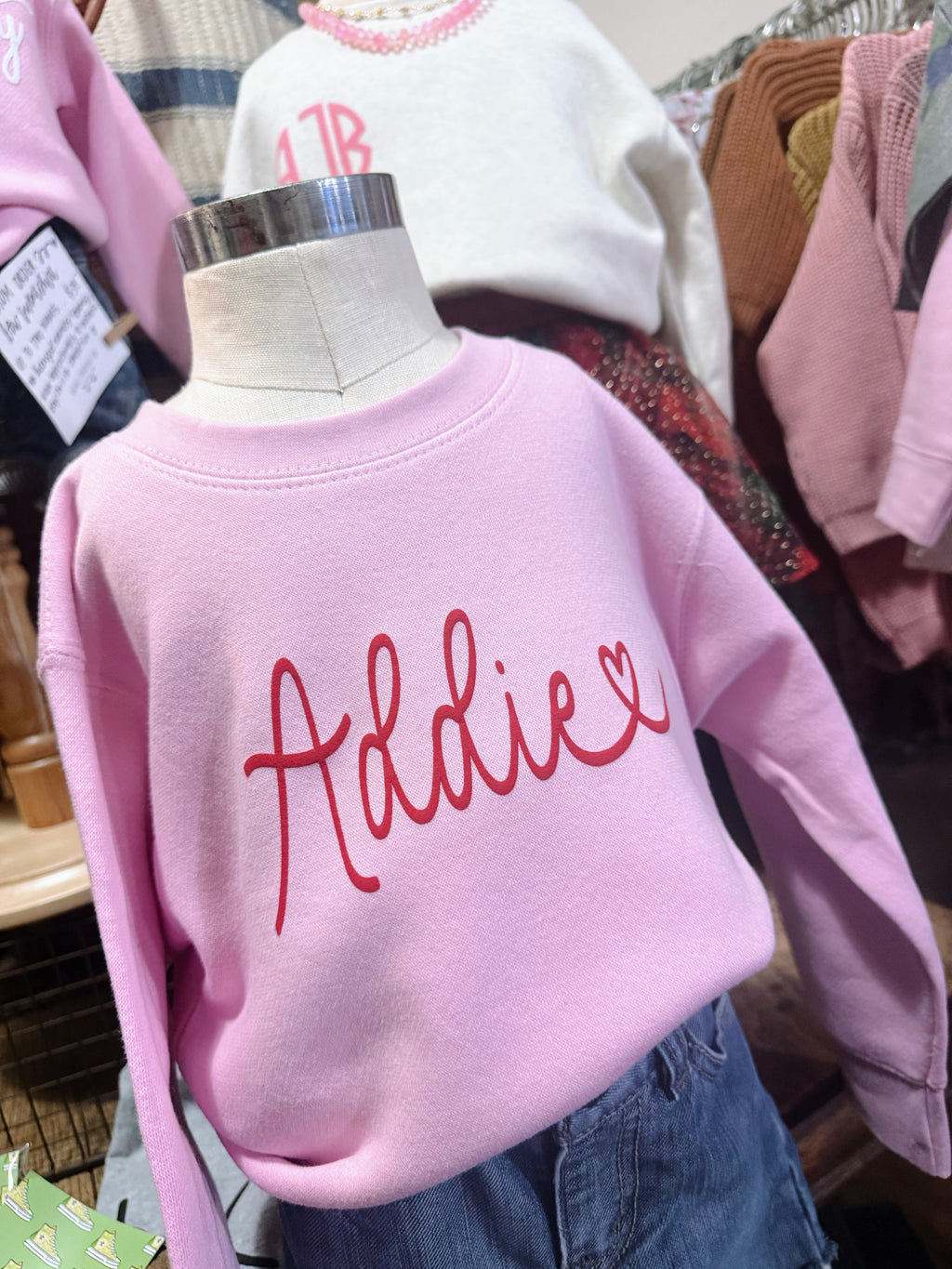 Pink sweatshirt with 'Addie' printed on it on a mannequin, surrounded by other clothing items.