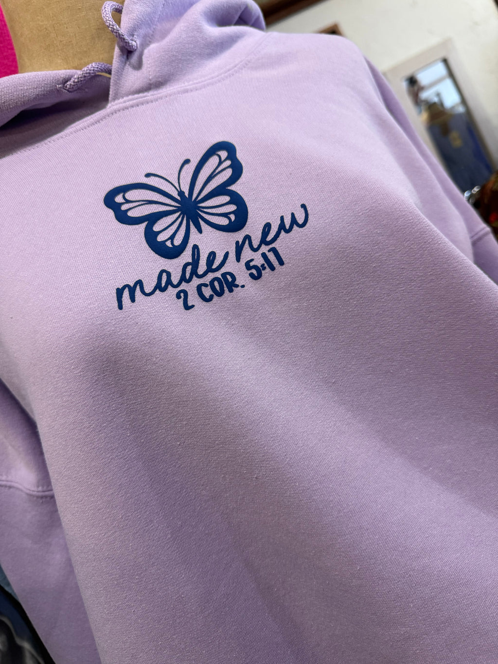 lilac colored hoodie on mannequin with blue butterfly print and words made new in cursive underneath, 2 COR. 5:17 under that. hoodie is shown on a mannequin with a mirror and other things blurred in the background