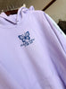 lilac colored hoodie with a blue butterfly logo and text on a hanger. other objects in background